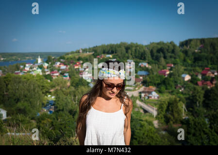 Junge Frau mit Sonnenbrille und Verband steht auf einem Hügel im Sommer Tag und blickte nach unten. Schöne gir im Park auf Hintergrund des kleinen Dorfes Stockfoto