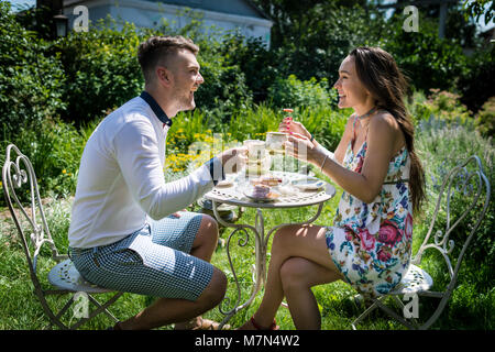 Junges Paar sitzt am Tisch und hält Tassen mit Tee im Garten. Zwei Menschen Lächeln und Lachen im Hof. Junge Frau mit Freund Tee trinken. Stockfoto