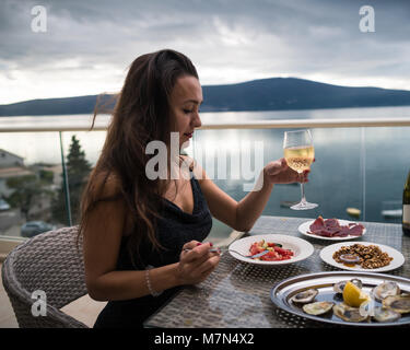 Junge nachdenkliche Frau sitzt auf dem Balkon am Tisch mit Austern, Fleisch und Salat und hält ein Glas Sekt. Mädchen ist bereit zu essen. Meer und Berge Stockfoto