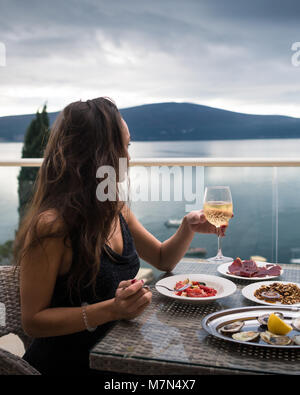 Junge Frau sitzt auf dem Balkon am Tisch mit Austern, Fleisch und Salat und hält ein Glas Sekt. Mädchen schauen auf das Meer und die Berge. Seitenansicht Stockfoto