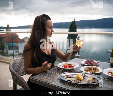 Junge Frau sitzt auf dem Balkon am Tisch mit lokalen Speisen und hält ein Glas Sekt. Mädchen schaut auf das Meer und die Berge. Seitenansicht Stockfoto