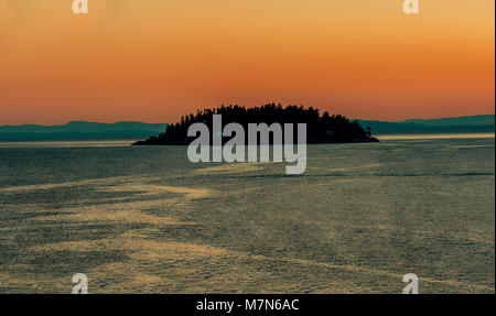 Eine Insel im Ozean mit schwarzen Silhouetten der Bäume und der leuchtenden Fenster der Häuser, vor dem Hintergrund der Berge und eine orange rosa Sonnenuntergang, Stockfoto