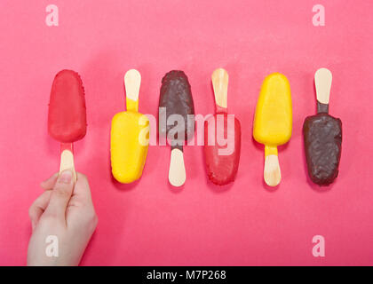 Weibliche Hand, die bunten Süßigkeiten, Kuchen Eis am Stiel auf einem hellen rosa Hintergrund, abwechselnd Aromen und die Richtung der Kuchen knallt. Fun Geburtstag pa Stockfoto