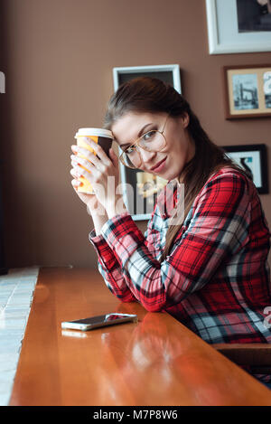 Ein Student Mädchen in einem Plaid Shirt und Brille, an einem Tag in einem Cafe an einem Tisch am Fenster, mit einer gelb braun Tasse Tee. Er hält einen heissen Kaffee in der Hand. Eine brünette Frau ist glücklich lächelnd, neben ihr ist ein Smartphone. Stockfoto