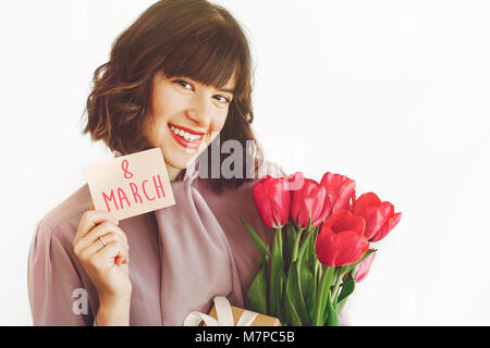 Der glückliche Frauen Tag Text auf der Grußkarte. 8. März. Gerne stilvolle Girl holding Card und rosa Tulpen und Geschenk, Lächeln auf weißem Hintergrund. Glückliche Frauen Stockfoto