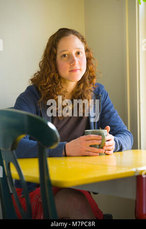 Porträt einer jungen Frau, die an einem Tisch sitzen mit ihren heißen Getränk in den Händen. Stockfoto