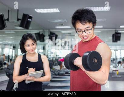 Hübscher junger Mann bei der Arbeit mit Muskeln Hantel mit schönen jungen weiblichen Trainer im Fitnessraum Stockfoto