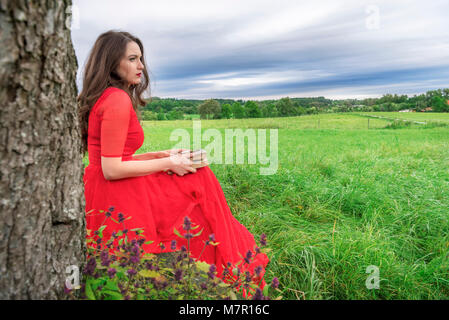 Schöne Frau, die in einem eleganten roten Kleid, sitzen auf einer Bank unter einem Baum, mit einem Buch und Taschenuhr in Ihren Händen, in Schwäbisch Hall, Deutschland. Stockfoto