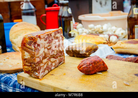 Headcheese und geräucherte Wurst auf einem hölzernen Schneidebrett der Outdoor Küche, Menschen im Hintergrund sind die Zubereitung von Fleisch für handgefertigte Würstchen. Stockfoto