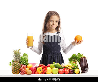 Mädchen, dass ein Glas Saft und ein orange hinter einem Tisch mit Obst und Gemüse auf weißem Hintergrund Stockfoto