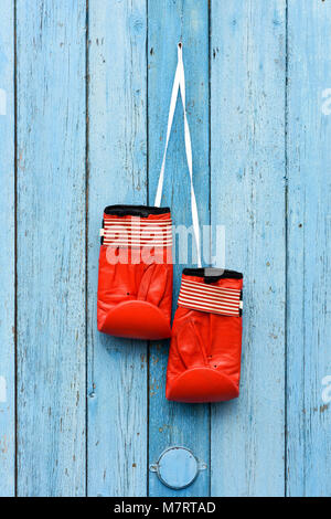 Ein Paar rote Leder Boxhandschuhe an einer weißen Kordel hängen, eine blaue alte, gesprungene Wand aus Holz Stockfoto