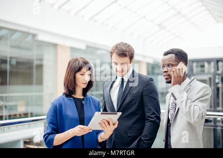Drei fröhliche multinationale Unternehmer stehen und mit Tablette im Büro zusammen Stockfoto