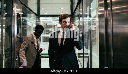 Zwei multinationalen Unternehmen Mann, der Aufzug und der Verwendung des Smartphones. Geschäftsleute in großen gläsernen Aufzug in modernen Büro Stockfoto