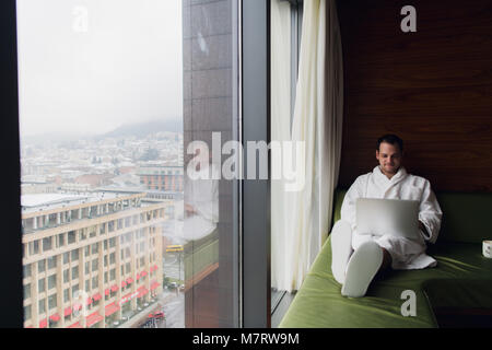 Unternehmer zu Hause oder in der Reise. Seitenansicht Portrait von hübscher junger Geschäftsmann sitzt auf dem Bett und mit Laptop. Panoramafenster mit schönen Sonnenaufgang Stadt Landschaft im Hintergrund Stockfoto