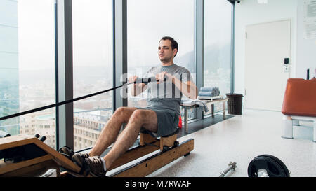 Rudern mit Strom. Seitenansicht des jungen Mannes in Sportkleidung, Rudern vor Fenster im Fitnessstudio. Stockfoto