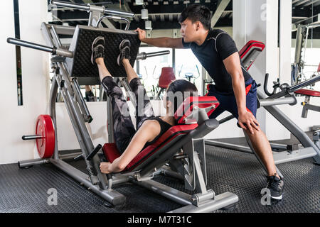 Sportliche brünette Frau mit Gewichte drücken Sie die Maschine für die Beine mit Hilfe Ihrer persönlichen Trainer im Fitnessstudio. Stockfoto