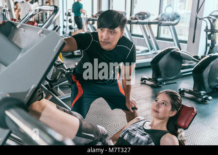 Personal Trainer hilft seinem weiblichen Kunden am Bein drücken sie arbeiten in der Turnhalle. Konzept der indoor Fitness gesunde Lebensweise. Stockfoto