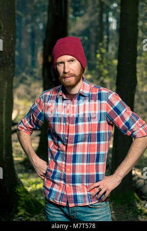 Portrait von ein ernster Mann mit Bart in einem Plaid Shirt und Hut im Wald Stockfoto