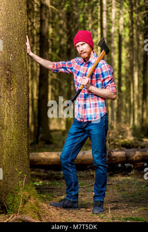 Vertikale Portrait eines bärtigen Holzfäller in voller Länge mit einer Axt in der Nähe von einem Baum im Wald Stockfoto
