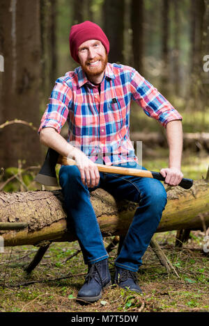 Ein lächelndes bärtigen Holzfäller in einen Hut und Hemd mit einer Axt sitzt auf einem Baumstamm im Wald Stockfoto