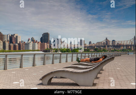 Man Sonnenbaden auf einer hölzernen Liegestuhl in der Gantry Plaza State Park über dem East River von Manhattan mit der Queensboro Bridge in der Ferne Stockfoto