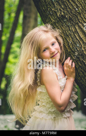 Kleines Mädchen mit langen blonden Haaren durch Baum im Wald Stockfoto
