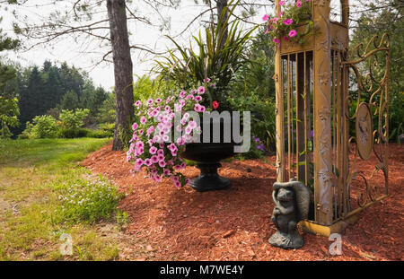 Nahaufnahme der Frosch Statue neben der Säule auf goldenen Schmiedeeisen metall Laube Tor eingerichtet mit rosa Petunien, schwarz Pflanzmaschine mit Petunia x hybrida Stockfoto