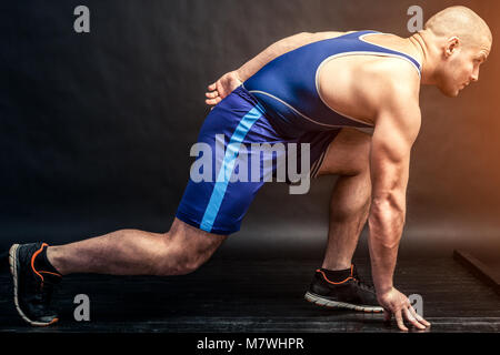 Ein junger sportlicher Mann in Blau wrestling tricky und blauen Shorts macht eine Ausdehnung auf einem schwarzen Hintergrund isoliert in einem Photo Studio Stockfoto