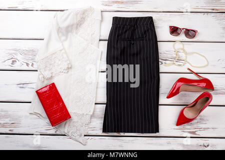 Woman's Zubehör flach auf weißem Holz Hintergrund. Gefaltete weisse Spitze Bluse mit schwarzen Hosen, Rote Mappe, glänzende Schuhe, Sonnenbrille und Halskette. Stockfoto