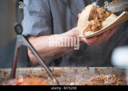 Mann im grauen Hemd, zog Schweinefleisch Street Food holding Sandwich mit Rinde und Zangen in den Vordergrund Stockfoto