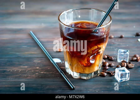 Kaffee mit Eis und Milch in ein Glas, Eiswürfel, Stücke von Schokolade und Plätzchen auf einem dunklen Holztisch gießen Milch in ein Glas. Low Key Stockfoto
