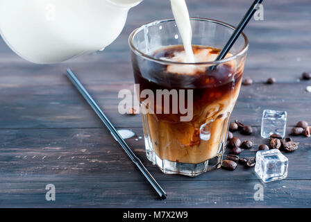 Kaffee mit Eis in ein Glas Milch, gegossen in ein Glas, Eiswürfel, Stücke von Schokolade und Plätzchen auf einem dunklen Holztisch Low Key Stockfoto