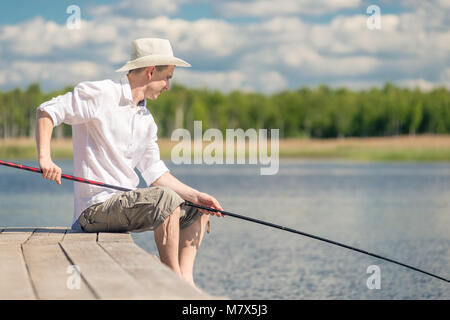 Männliche Fischer mit Angel sitzt auf dem Pier in der Nähe des Sees Stockfoto