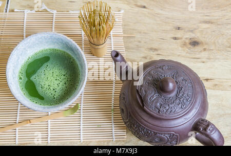 Oben Blick auf Japanische Tee Zeremonie Elemente einschließlich einen Stein Schüssel mit Matcha grüner Tee, Bambusbesen, Schaufel, und einem braunen Asiatische Teekanne auf Bambus Matte. Stockfoto