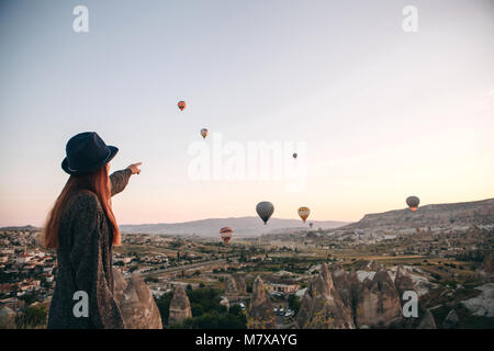 Ein Tourist girl in einen Hut bewundert Heißluftballone fliegen in den Himmel über Kappadokien in der Türkei. Beeindruckender Anblick. Tourismus Stockfoto