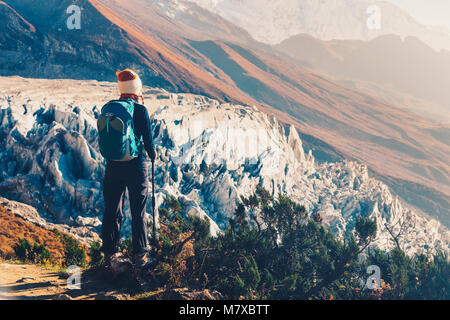 Stehende junge Frau mit Rucksack auf dem Gipfel gegen die schönen Berge und Gletscher bei Sonnenuntergang. Landschaft mit Mädchen, Felsen mit schneebedeckten Gipfeln Stockfoto