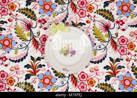 Eine Blume, die Platte auf einem geblümten Stoff mit einem gelben Schmetterling am Rande versteckt. Camouflage Spiel. Minimale Farbe still life Fotografie. Stockfoto