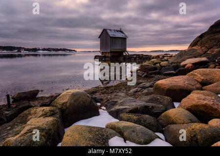 Verschneite Landschaft Blick auf die felsige Küste und ein entfernter Kabine bei bewölktem Himmel, an der Westküste von Göteborg, Schweden. Stockfoto