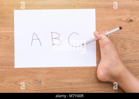 Wort 'ABC' aus weißem Papier schreiben, indem Sie mit der rechten Fuß von behinderten Menschen auf hölzernen Schreibtisch. Verwenden Sie für die Spende für Behinderte Konzept. Fokus auf Fuß Top View Stockfoto