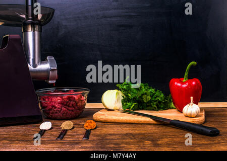 Küche noch durch einen elektrischen Fleischwolf, Gewürze in Löffel, Petersilie, Zwiebel, Knoblauch und Paprika mit einem Messer auf einem Holztisch auf dem Hintergrund o Stockfoto