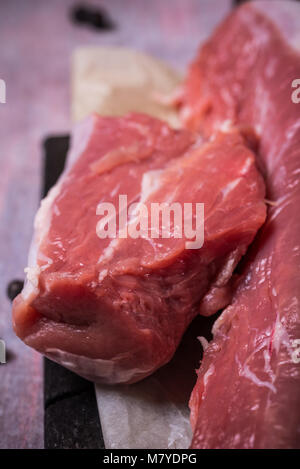 Vertikale Foto mit Details der ganzen Schweinefilet. Rohes Fleisch ist auf dunklen vintage Holzbrett und ein Stück Papier. Paar Wacholderbeeren sind Spille Stockfoto