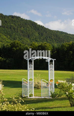 Weiße schmiedeeiserne Metall Laube Tor in der Mitte der gepflegten grünen Rasen im privaten Hinterhof Land Garten im Sommer Stockfoto