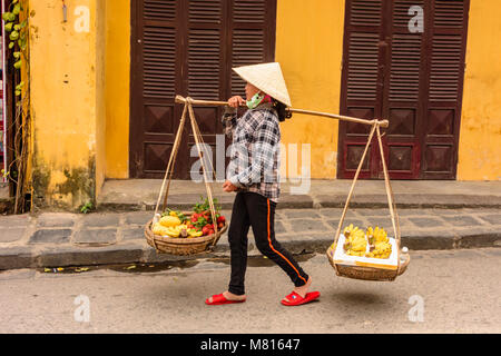 Eine vietnamesische Frau das Tragen eines traditionellen Bambus konische Hut trägt Früchte für den Verkauf in Körbe aus dem Bambus Durchführung Pol in Hoi An, Vietnam Stockfoto