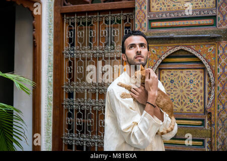 Junge muslimische Mann in traditioneller Kleidung, die eine Gelbe Katze im traditionellen marokkanischen Ambiente vor einem geschmückten Wand Stockfoto