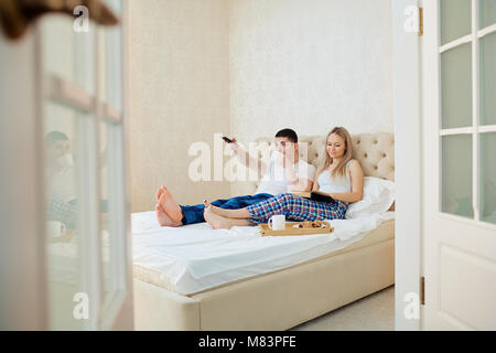 Ein paar im Bett frühstücken, fernsehen, lesen ein Buch im Schlafzimmer. Stockfoto