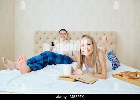 Ein paar im Bett frühstücken, fernsehen, lesen ein Buch im Schlafzimmer. Stockfoto