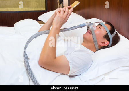 Mittleres Alter asiatischer Mann in seinem Bett tragen CPAP-Maske und mit seinem Smart Phone in der Nacht Stockfoto