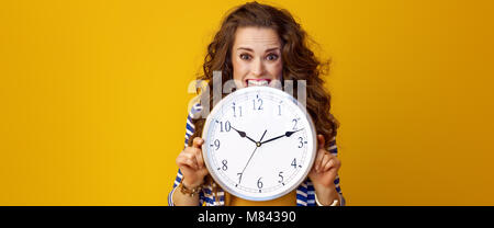 Trendy Frau in gestreifte Jacke auf gelben Hintergrund beißen Clock betonte Stockfoto