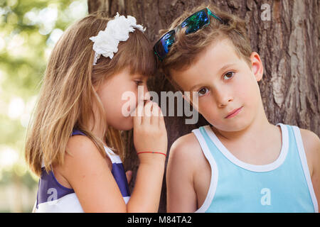 Erste Liebe. Kleine süße Junge und glückliche Mädchen spielen im Park und Spaß haben. Mädchen flüstert ein Geheimnis in einen jungen Ohr Stockfoto