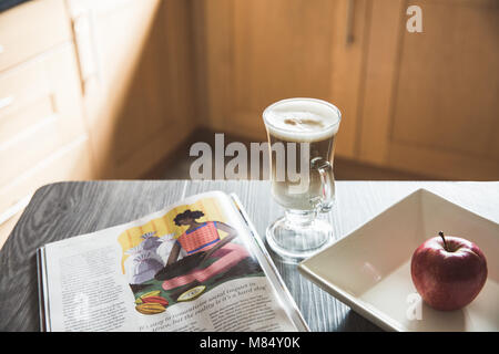 Ein Frühstückstisch mit einem hohen Glas Macchiato oder Cappuccino und einer Zeitschrift in einem Interieur zu lesen. Stockfoto